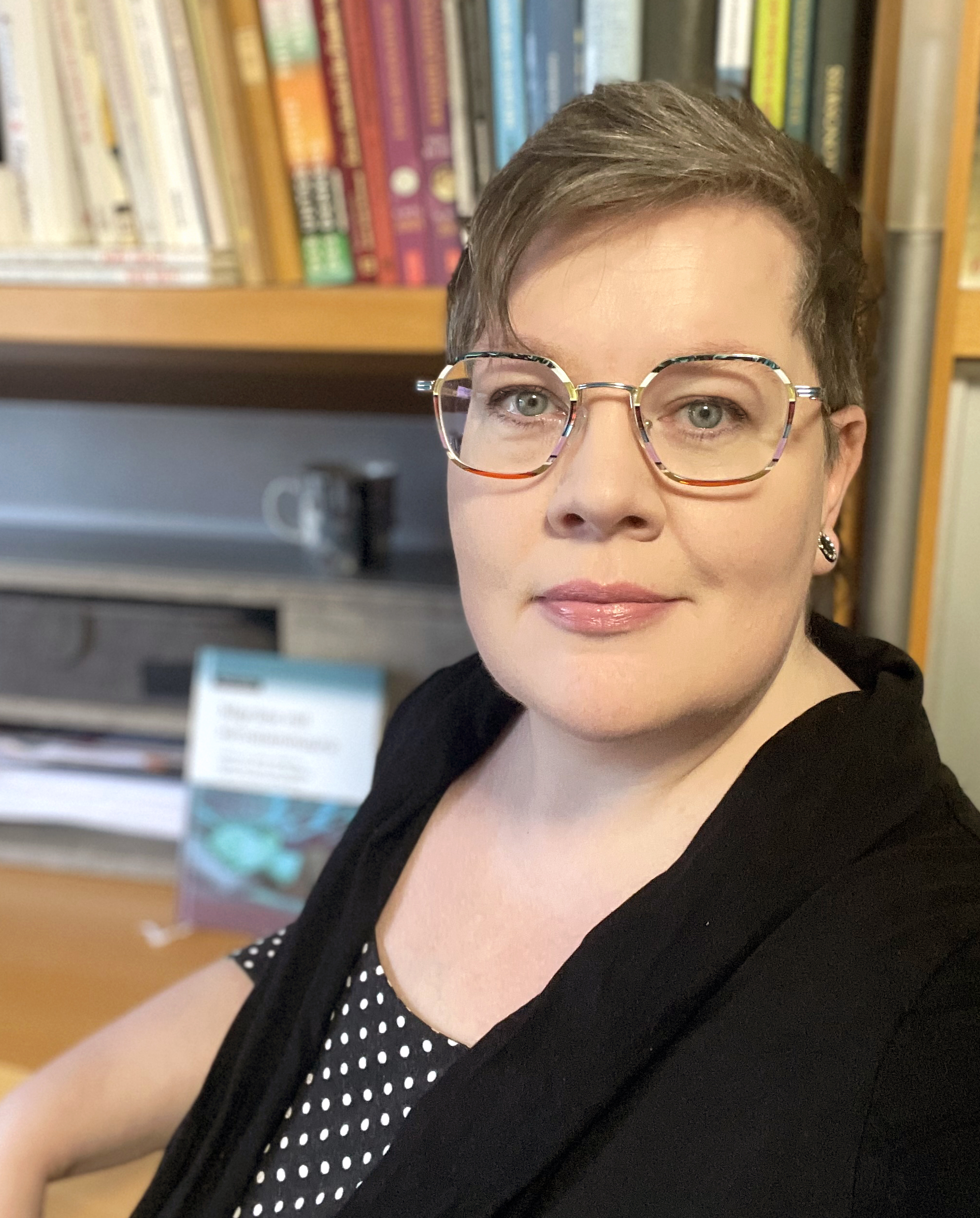 Foto der Seitenbetreiberin. Eine Frau mit Brille und Kurzhaarfrisur. Sie trägt ein schwarzes Oberteil. Im Hintergrund sieht man ein Bücherregal und etwas verschwommen ein Buch der Autorin.